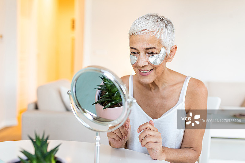微笑的年长女士在脸上敷焕活眼膜。老年女性喜欢在镜子前做面部按摩、去角质，并在眼睛下敷上面膜图片素材