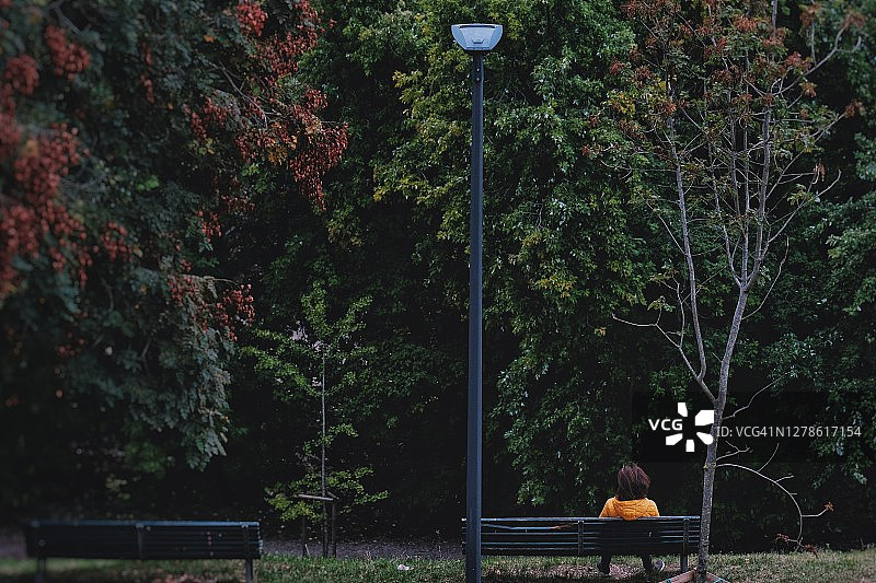穿黄色雨衣的孤独男人背对着坐在日落黑暗的森林长椅上图片素材