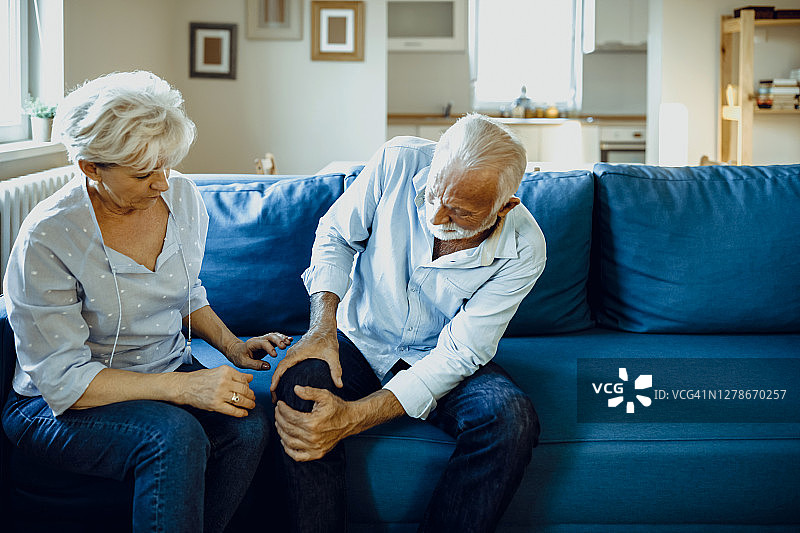 老年夫妇在家中，英俊的老人膝盖酸痛，他迷人的老伴扶着他图片素材