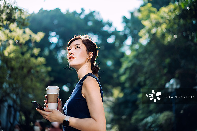 自信的年轻亚洲女商人漫步在城市公园，一边使用手机，一边手持咖啡杯图片素材