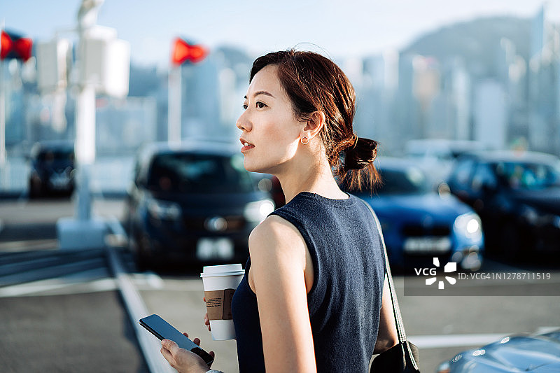 自信且有抱负的亚洲女商人手持智能手机和咖啡，在户外停车场仰望城市天际线图片素材