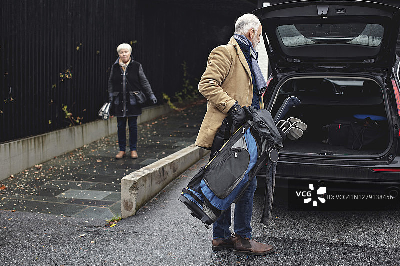 年长的男人拿着高尔夫球包站在汽车后备箱旁，女性伴侣在冬季站在人行道上图片素材