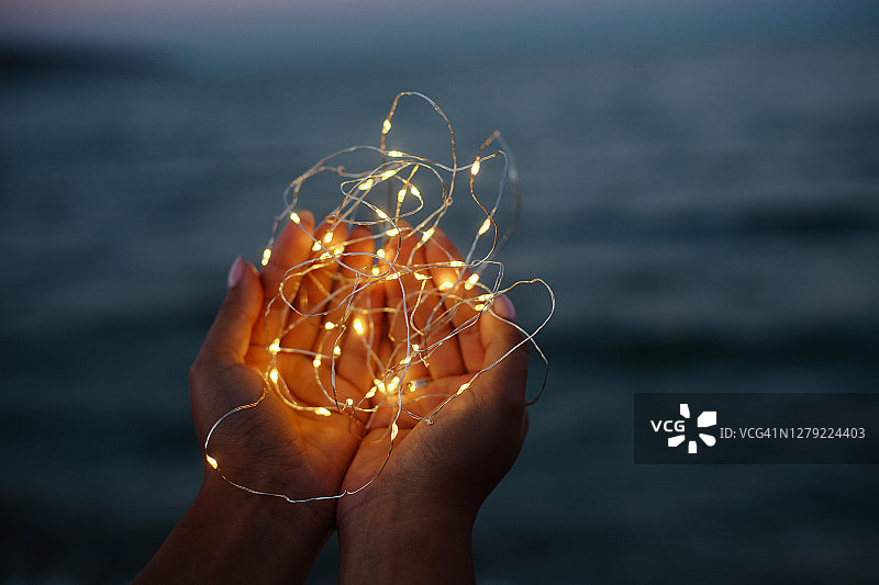 蓝色大海背景下，女性手中拿着一串串灯，浪漫的户外活动、节日或假日照片图片素材