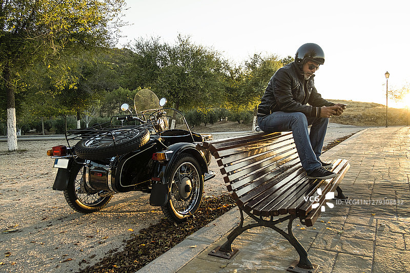 中年男人在长椅上，背景是边三轮摩托车，用手机发送短信，旅途中休息图片素材