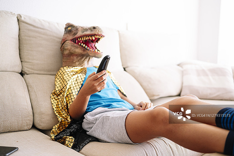 男孩戴着恐龙面具，在家里的沙发上放松看电视图片素材