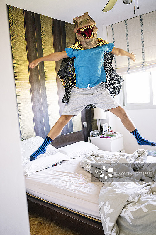 男孩戴着恐龙面具和披风在家里的床上跳跃图片素材