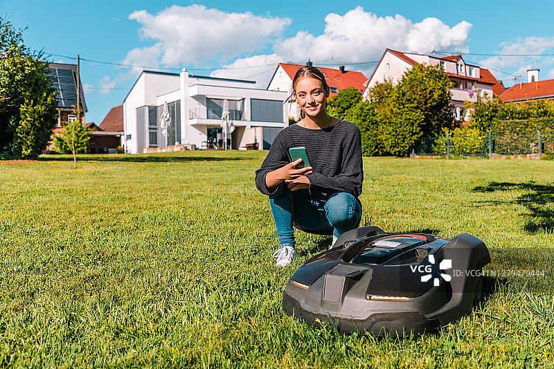 微笑的女人用手机App控制机器人割草机图片素材