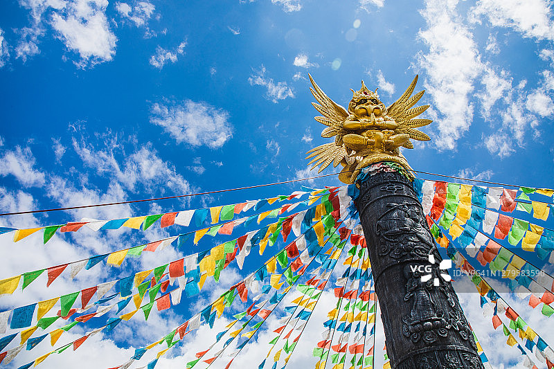 金翅鸟坐在图腾柱上，背景是藏传佛教祈祷旗和蓝天图片素材
