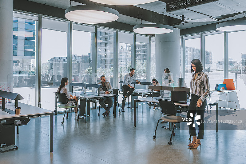 在疫情期间，商务同事团队在现代办公室中通过保持社交距离在亚克力玻璃后面工作图片素材