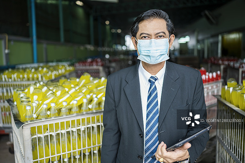 一家果汁饮料厂的成功的成熟亚洲男性老板的肖像。站在生产产品过程中。在covid-19爆发期间，他戴着医用口罩。图片素材