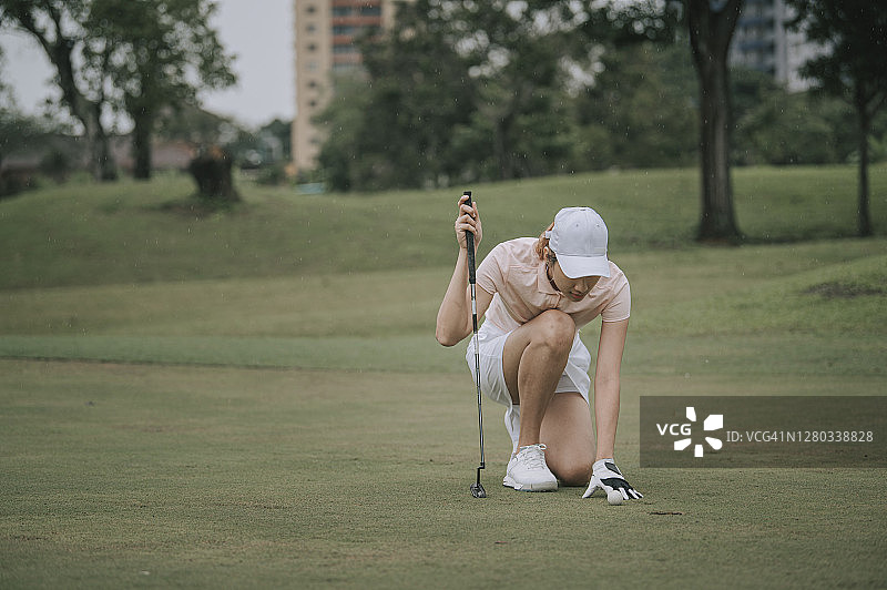 亚洲华裔女高尔夫球手戴着白色手套跪地测量高尔夫球和推杆果岭球洞之间的距离图片素材