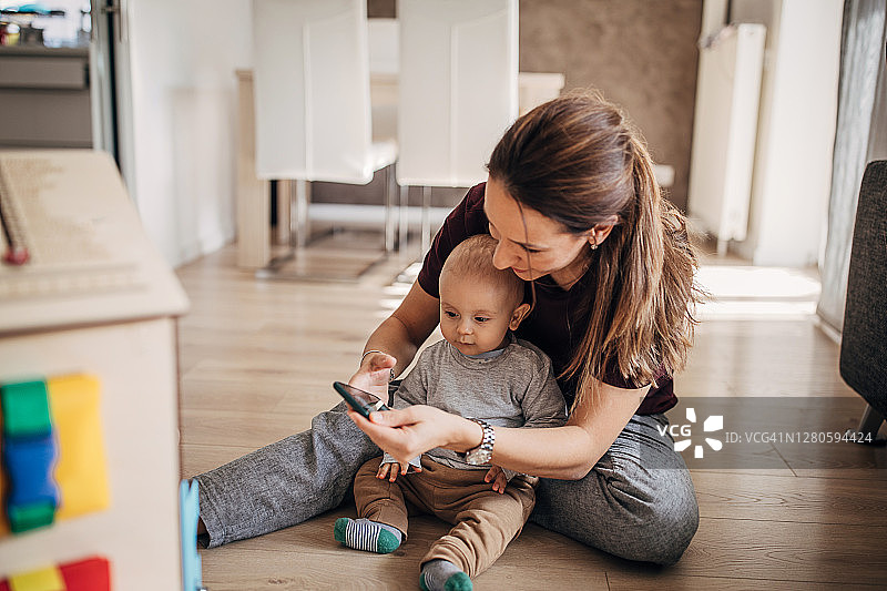 年轻妈妈和宝宝儿子坐在地板上，使用智能手机图片素材