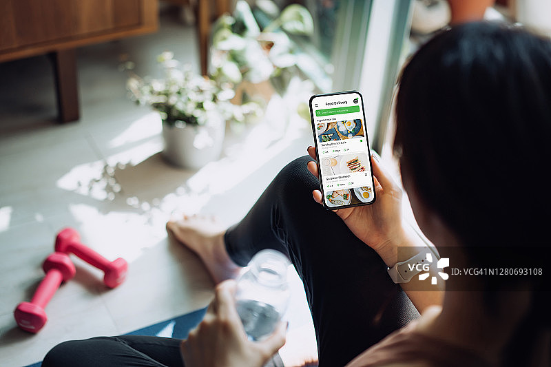 肩后视角：运动后亚洲女性使用手机订购健康食品图片素材