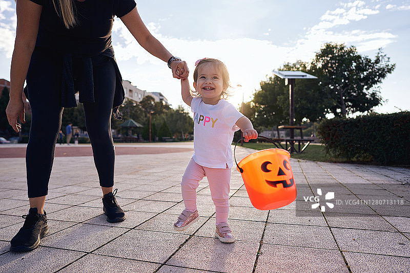 兴奋的女婴拿着桶要糖果，母亲帮助她走路图片素材