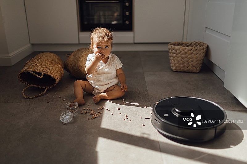 可爱的米色衣服女宝宝与现代扫地机器人在瓷砖地板上图片素材