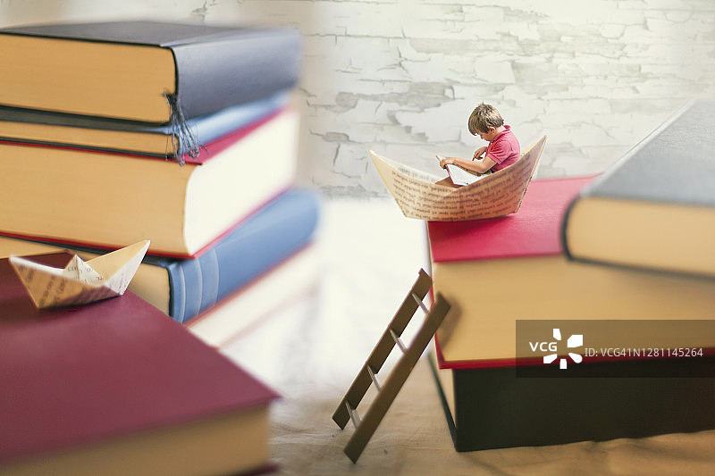 男孩在一艘小纸船里读书，纸船在一堆巨型书本上图片素材
