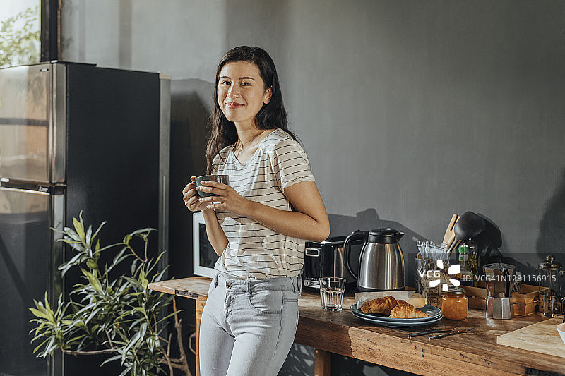 快乐的亚洲女人在厨房站立喝茶图片素材