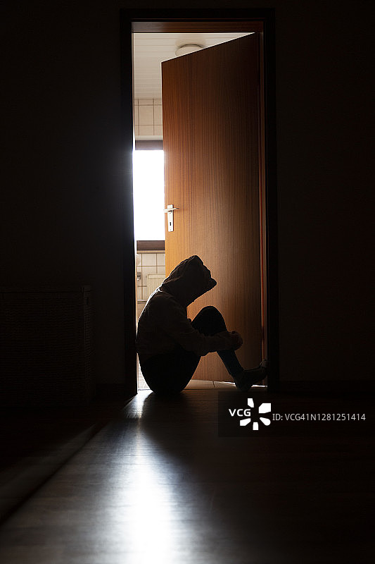 孤独的青少年穿着连帽上衣坐在门口的黑暗框架中，被身后充满阳光的房间照亮图片素材