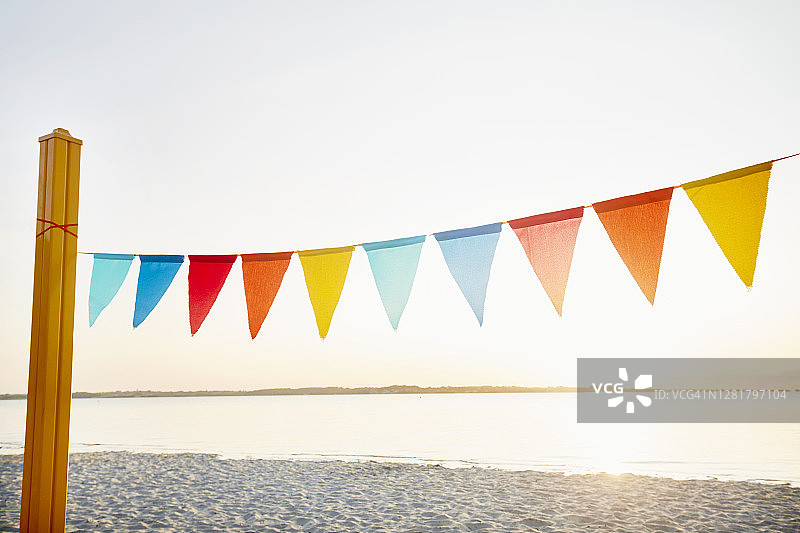 挂在海滩黄色杆子上，以天空和太阳为背景的派对装饰彩色三角旗／旗帜链图片素材