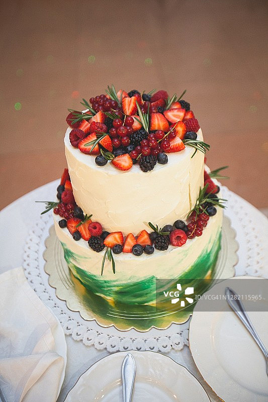 白色木桌上装饰着浆果的优雅双层婚礼蛋糕，附近有盘子和餐具图片素材