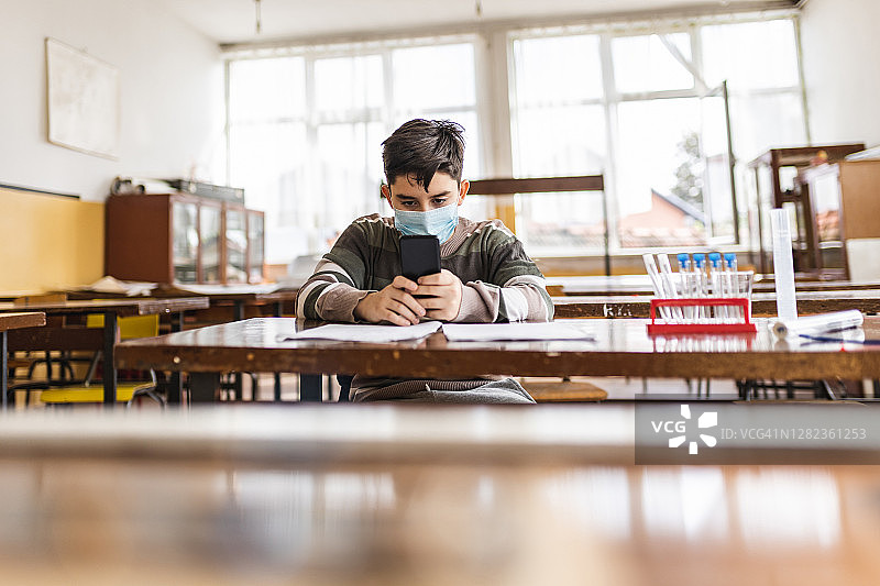 小学生戴着防护口罩坐在学校课桌旁使用手机图片素材