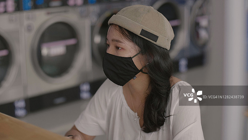 在自助洗衣店等待洗衣的戴面罩的年轻女子图片素材