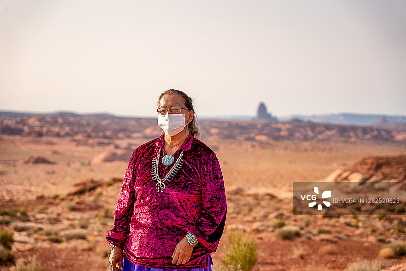 一位美国原住民纳瓦霍族妇女的肖像，身着民族服装和绿松石珠宝，戴着面具，背景是亚利桑那州卡延塔附近的船岩纪念碑图片素材