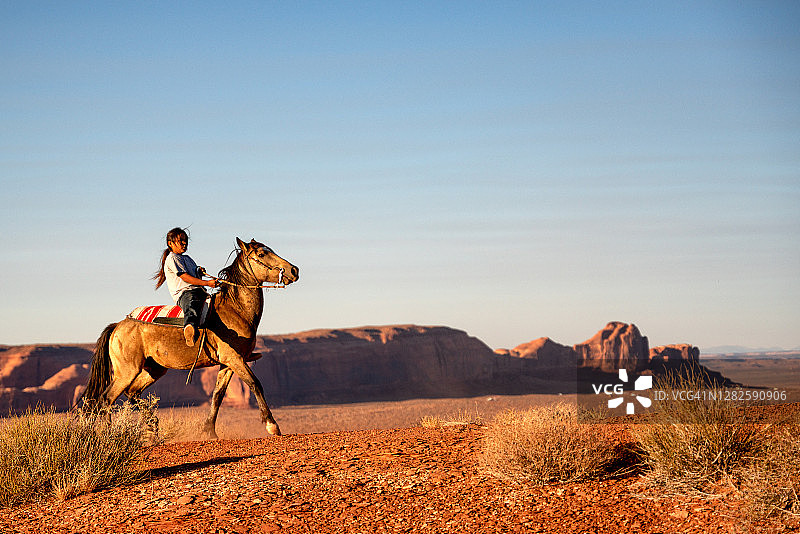 一位年轻的纳瓦霍男孩在纪念碑谷的家中高原上嬉戏地骑着马图片素材
