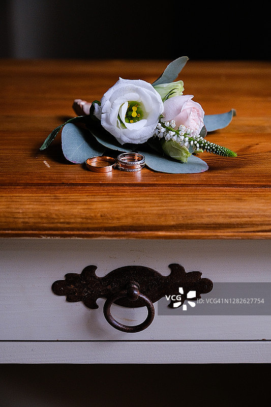 新娘和新郎的结婚戒指在复古木制抽屉柜、桌子或床头柜上。旁边是一朵花或胸花。图片素材