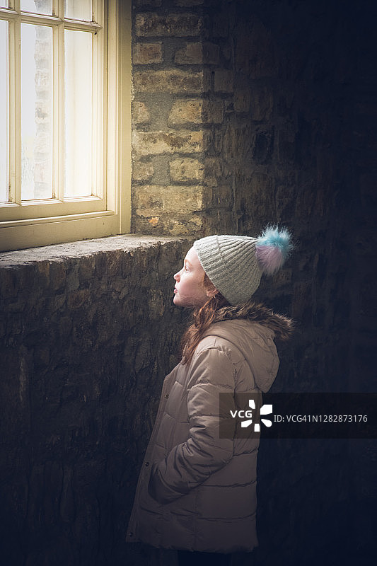 年轻女孩望向窗外，昏暗的砖墙房间图片素材