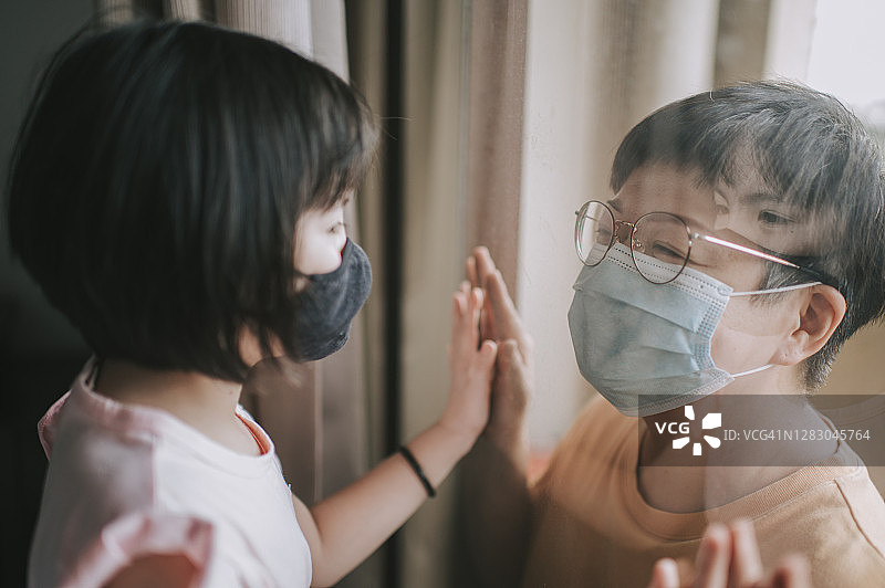 中国小女孩在屋内隔着窗户，戴着口罩看着她的母亲图片素材