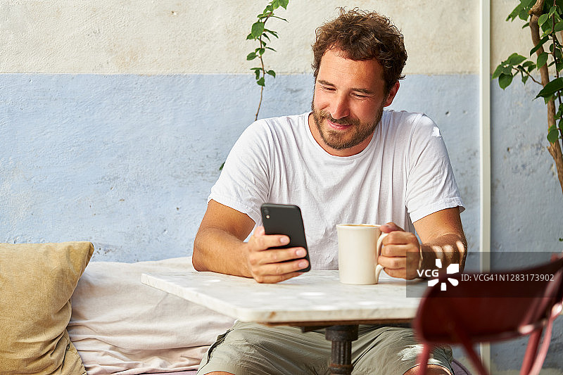 一位留着胡子的男人坐在咖啡店的桌旁，手里拿着咖啡杯，看着他的智能手机或iphone，面带微笑图片素材