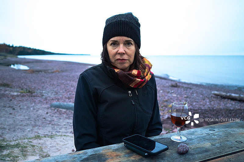 女人坐在岩石海滩的野餐桌旁，手持鸡尾酒和手机图片素材