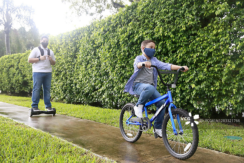 两名Z世代拉丁裔兄弟在热带环境中穿着休闲服装和防护口罩，骑着自行车和悬浮滑板在人行道上行驶图片素材