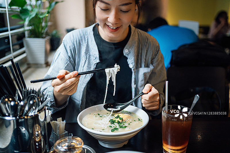 微笑的年轻亚洲女性坐在窗边，享用一碗新鲜的当地风味鸡肉汤面和一杯港式冰镇柠檬茶图片素材