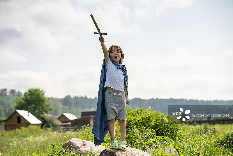 穿着披风，拿着玩具剑，站在岩石上对着天空的顽皮男孩图片素材