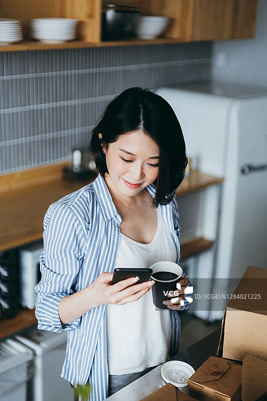 快乐的年轻亚洲女性查看手机，确认收到的快递，手里拿着咖啡图片素材