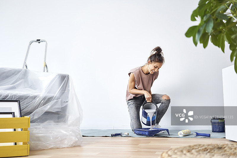 微笑的年轻女子跪在家里，将油漆倒入漆盘图片素材