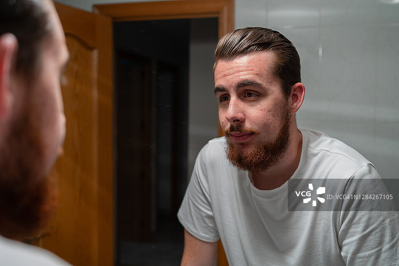 在浴室镜子前看自己的年轻红发胡须痤疮男孩图片素材