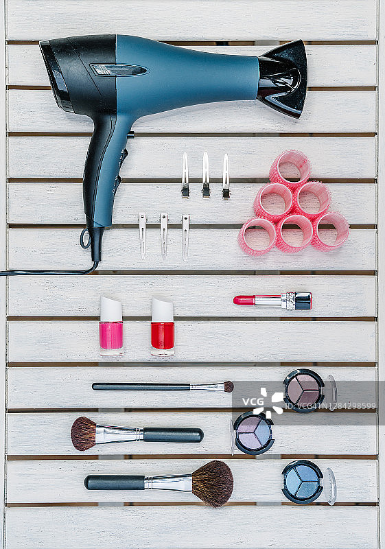 化妆台上的一套化妆品特写：吹风机、粉色卷发器、金属夹、口红、指甲油、面刷和彩色眼影，竖版照片图片素材