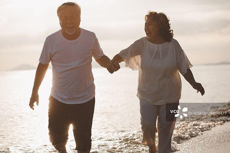 浪漫的亚洲老年夫妇手牵手在夏日海滩日落漫步，享受退休后的休闲旅行和活动。图片素材