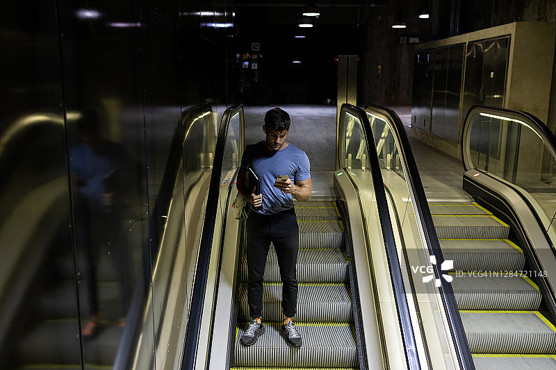 在地铁自动扶梯上使用智能手机并手持平板电脑的年轻男人图片素材