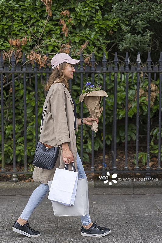 年轻女子在人行道上拿着购物袋和鲜花走路图片素材