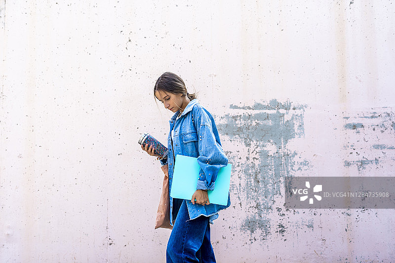 阳光明媚的日子里，靠墙拿着书和瓶子行走的少女图片素材