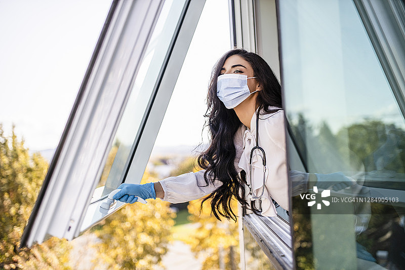 新冠疫情期间，年轻女医生戴着医用口罩和外科手套，在办公室里望着窗外图片素材