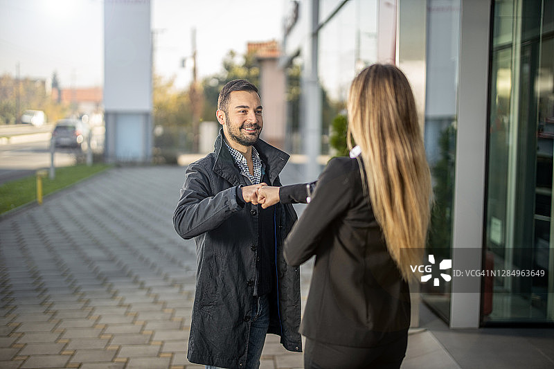 年轻男人到达汽车经销店，女销售员在入口处欢迎他图片素材