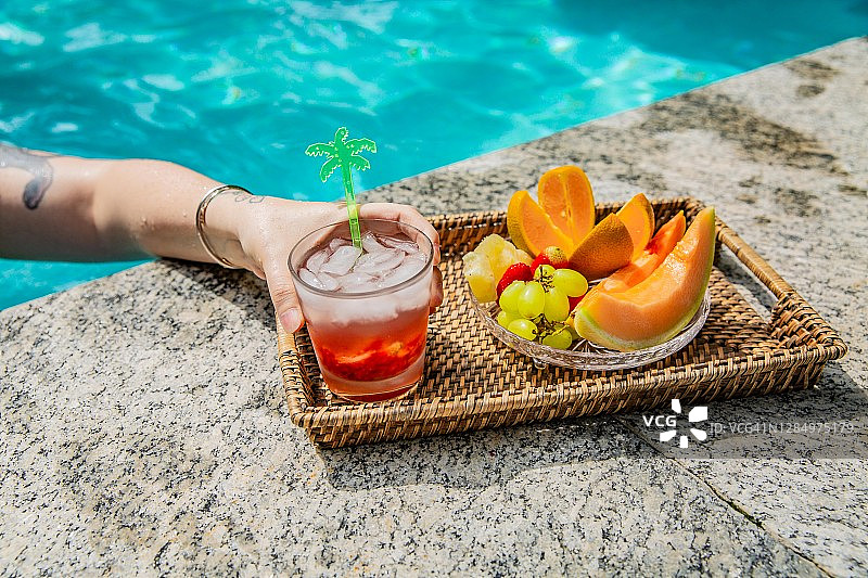 阳光明媚的夏日，女子在泳池边吃水果、喝凯匹林纳鸡尾酒图片素材