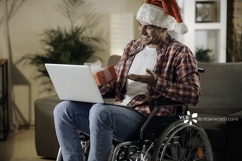坐在轮椅上，头戴圣诞帽的快乐年轻男人通过视频通话祝圣诞快乐图片素材
