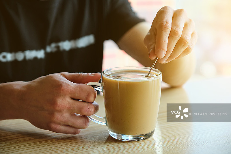 男子在咖啡馆木桌背景下手持一杯热咖啡或饮品，搅拌并饮用，早餐，咖啡休息图片素材
