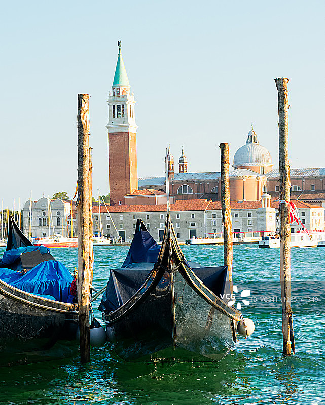 两艘意大利贡多拉船在码头的一些柱子上系着，漂浮在日落时分的蓝色海面上。后面是威尼斯的圣乔治马焦雷岛图片素材